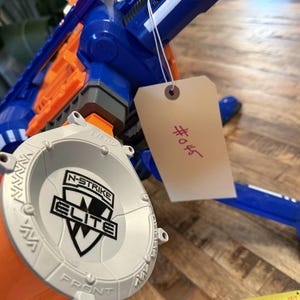 May include: A blue and orange toy gun with a gray drum magazine. The magazine is labelled "N-STRIKE ELITE". A tag with the number "#045" is attached. The toy gun is on a wooden surface.