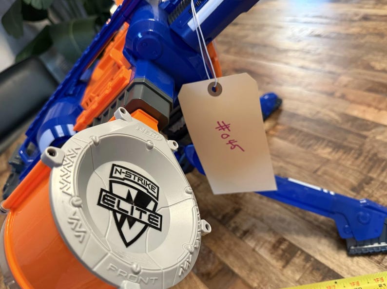 May include: A blue and orange toy gun with a gray drum magazine. The magazine is labelled "N-STRIKE ELITE". A tag with the number "#045" is attached. The toy gun is on a wooden surface.