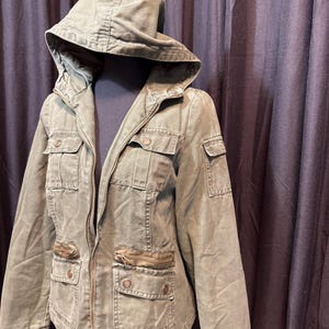 May include: Olive green hooded jacket with multiple front pockets and a zipper closure. The jacket is displayed on a mannequin, showcasing its design and fit. The jacket has a drawstring hood and a classic military-inspired style.