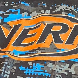 May include: Black t-shirt with the orange and black "NERF" logo and a blue and grey pixelated camouflage design. The text "&copy; 2020 Hasbro" is below the logo. A great shirt for fans of the NERF brand.
