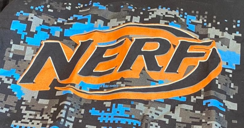 May include: Black t-shirt with the orange and black "NERF" logo and a blue and grey pixelated camouflage design. The text "&copy; 2020 Hasbro" is below the logo. A great shirt for fans of the NERF brand.