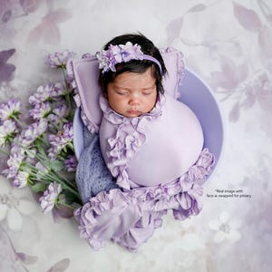 May include: A newborn infant wrapped in a light purple swaddle with ruffled edges, resting in a matching purple bowl. The infant wears a purple floral headband. Beside the bowl are sprigs of light purple flowers. The text reads: Real image with face ai swapped for privacy.