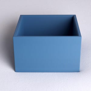 May include: A square, open-top box in a solid, muted blue color. The box has a simple, modern design with clean lines and a matte finish. The box is sitting on a white surface.