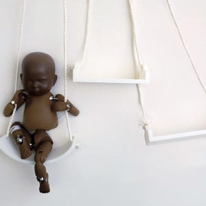 May include: A brown articulated doll sits on a white swing suspended by white rope against a white wall. Two additional white swings are also suspended from the wall. The doll has its eyes closed and is in a seated position.