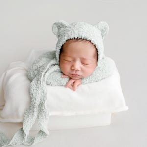May include: A newborn baby wearing a light green bear-eared bonnet and matching wrap, resting on a white pillow. The baby is peacefully asleep, with hands gently placed on the pillow. The soft, textured fabric adds a cozy feel.