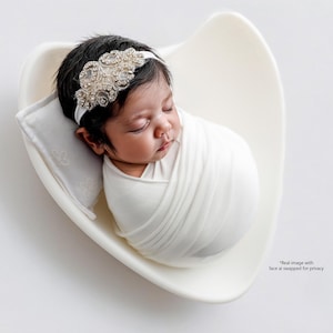 May include: A newborn infant with dark hair is wrapped in a white swaddle and resting in a white heart-shaped bowl. The infant wears a white headband featuring intricate silver and clear crystal embellishments, and rests on a small white pillow.
