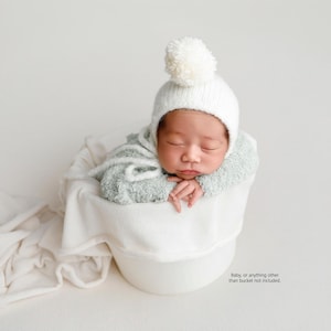 May include: A newborn baby wearing a white knit hat with a pom-pom and a light green hooded outfit, resting peacefully in a white bucket. A white blanket is draped over the bucket's edge.