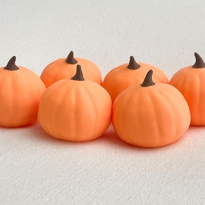 May include: A set of six small, round, orange pumpkins with smooth surfaces and dark brown stems. Each pumpkin features subtle vertical ridges and a rounded shape, arranged in a cluster on a plain, light-colored surface without any additional decor.