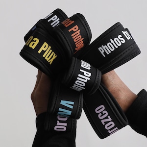 Könnte beinhalten: Eine Ansammlung schwarzer Armbänder mit farbigen Schriftzügen. Die Texte umfassen "Photos by", "na Plux" und weitere Namen. Die Armbänder werden zusammengehalten und sind aus Stoff.