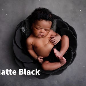 May include: A newborn baby with dark skin and curly hair rests inside a matte black, flower-shaped bowl with layered petals. The baby is wearing a small black headband. The text "Matte Black" is printed in white in the bottom left corner.