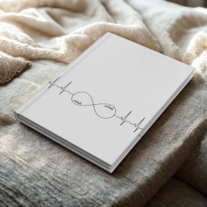 Puede incluir: Un diario blanco con las palabras "inhale" y "exhale" dentro de un símbolo de infinito, con un diseño de línea de latido cardíaco. El diario está sobre una tela de color neutro.