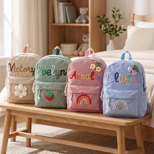 May include: Four corduroy backpacks in beige, sage green, dusty pink, and light blue. Each bag features a personalized name embroidered in cursive, along with a unique fabric applique: a white flower, a red strawberry, a rainbow, and flowers.