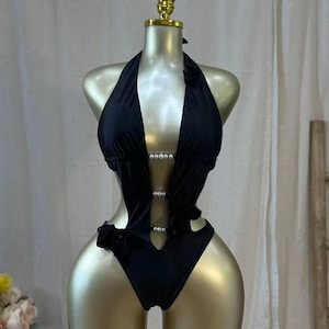 May include: Black halter-neck swimsuit with pearl accents and cut-out details. The one-piece design features a plunging neckline and a high-cut leg. The swimsuit is displayed on a gold mannequin, showcasing its form-fitting silhouette.