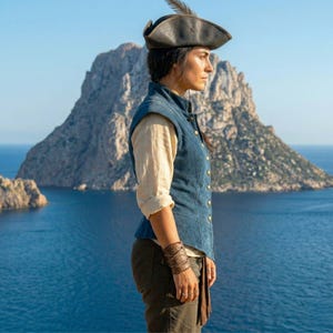 May include: A person wearing a dark leather tricorn hat with a feather, a blue vest, and a cream-colored shirt. The person is standing on a cliff overlooking the ocean, with a large rock formation in the background.