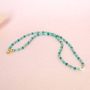 May include: A beaded necklace with turquoise, teal, and gold-colored beads. The necklace has a gold-colored clasp and a small white flower pendant at the bottom. The necklace is displayed on a light pink background.