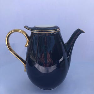 May include: A dark blue ceramic teapot with a gold handle and trim. The teapot has a lid with a white center and gold trim. The spout is curved, and the handle is a curved, open design. The teapot is likely for serving tea.