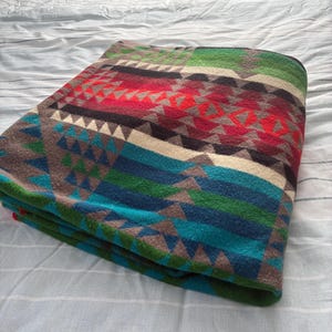 May include: Folded blanket with a geometric pattern in shades of red, green, blue, and gray. The blanket is folded neatly on a bed with a white and blue striped sheet. The blanket's design features triangles and other shapes.