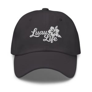 May include: A dark grey baseball cap featuring the embroidered words "Luau Life" in white script, accompanied by a white hibiscus flower. The cap has a curved peak and a button on the crown.