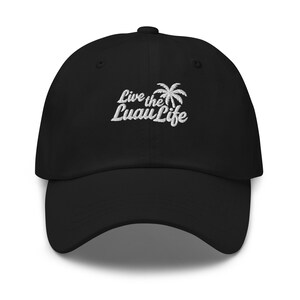 May include: Black baseball cap with a curved peak. The cap has white embroidered text and a palm tree graphic that reads "Live the Luau Life". The hat is a classic, adjustable style.