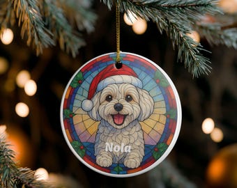 Puede incluir: Adorno navideño redondo con un perro blanco sonriente con un gorro de Papá Noel. El perro está en un diseño de vidriera con acentos de acebo. El nombre "Nola" está escrito debajo.