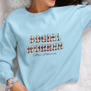 May include: Light blue sweatshirt with the words "SOCIAL WORKER" in a retro font with red, green, and white accents. Below the text is "Mrs. Petterson" in a script font. The sweatshirt is a crew neck style.