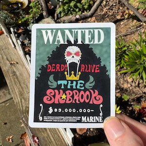 May include: A rectangular sticker featuring a Wanted poster design with a dark green background. It displays a white skull with pink heart-shaped sunglasses, a yellow crown, and the text "WANTED", "DEAD OR ALIVE", "THE SK BROOK", and "83,000,000-".