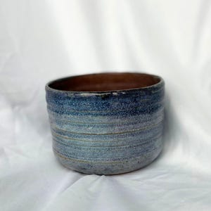 May include: A handmade ceramic bowl with a textured, horizontal striped design. The bowl features a gradient of blue hues on the exterior, transitioning to a brown interior. The bowl is likely used for serving food or as a decorative piece.