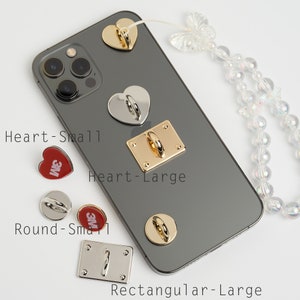 May include: A close-up of a black iPhone with a variety of phone ring holders attached to the back. The holders are in the shapes of hearts, circles, and rectangles. The phone is on a white surface with a beaded chain.