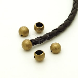May include: Five antique gold-toned metal beads with a hole through the center. The beads are round and have a slightly textured surface. A brown leather cord is visible in the background.