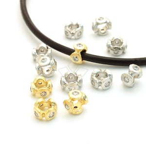 May include: A collection of silver and gold metal beads with clear round stones. The beads are arranged on a white surface with a brown leather cord.