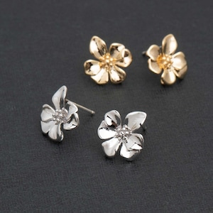 May include: Four flower-shaped stud earrings. Two are gold-colored, and two are silver-colored. The earrings have a detailed petal design and are set against a dark background.