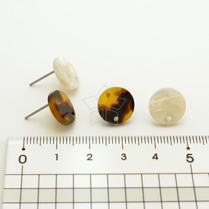 May include: Three pairs of earrings, each with a round, flat, decorative piece. One pair is brown and black, one is white, and one is a mix of white and brown. The earrings are shown on a ruler with markings from 0 to 5.
