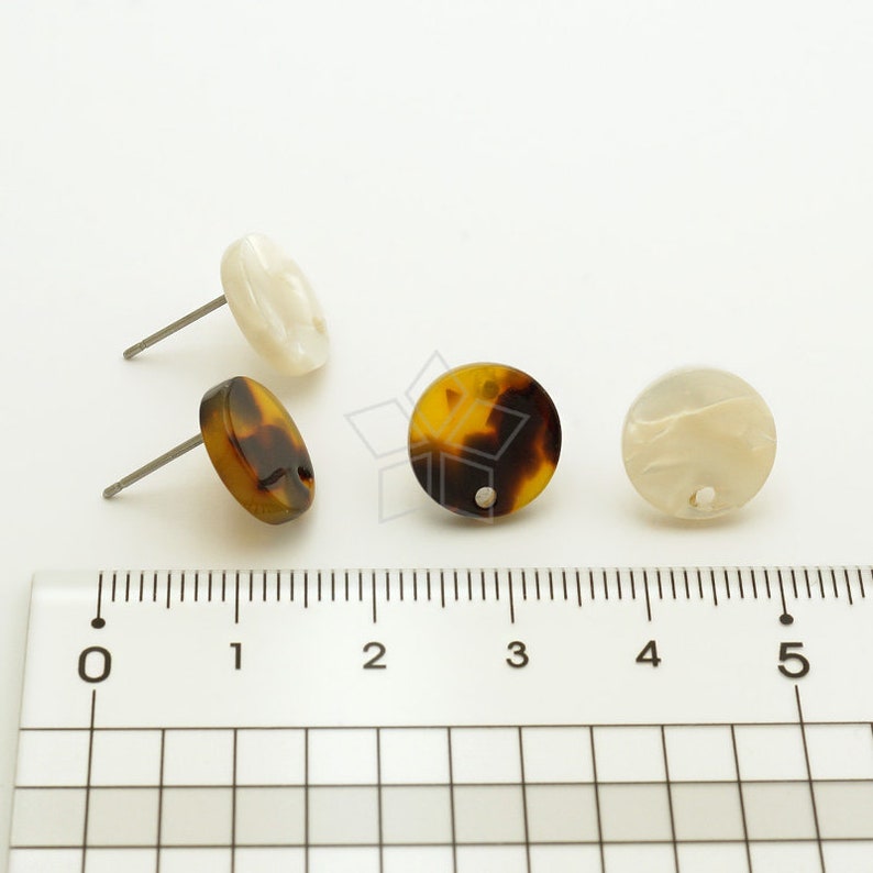 May include: Three pairs of earrings, each with a round, flat, decorative piece. One pair is brown and black, one is white, and one is a mix of white and brown. The earrings are shown on a ruler with markings from 0 to 5.