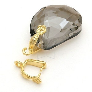 Pode incluir: Um pingente de cristal facetado em forma de lágrima, cinza esfumaçado, com uma argola decorativa dourada. Um fecho dourado separado também é visível. A argola e o fecho apresentam padrões florais e de folhas.