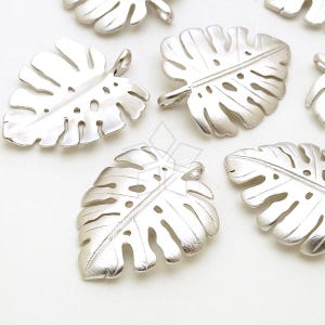 May include: Silver-toned metal charms shaped like monstera leaves. The leaves have a cut-out design and are perfect for jewelry making.