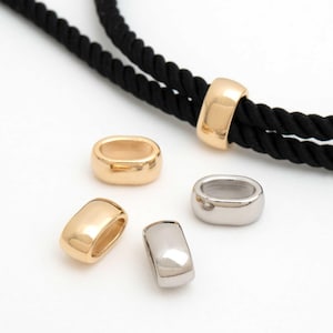 Op de afbeelding: Goud- en zilverkleurige ovale metalen kralen. De kralen worden getoond met een zwart touw. De kralen zijn glad en glanzend, met een afgeronde rechthoekige vorm. De kralen worden waarschijnlijk gebruikt voor het maken van sieraden of als decoratieve elementen.