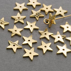 May include: A collection of gold-toned metal star-shaped charms with a hole in the center. The charms are scattered on a gray surface.