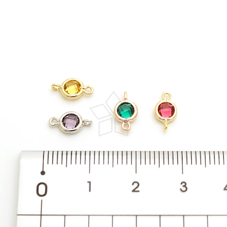 May include: Four colorful gemstone connectors with gold and silver-tone settings. The gemstones are yellow, purple, green, and red. Each connector has a loop on either side for attaching to other jewelry components. A ruler is in the foreground.