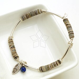 May include: Silver bracelet with a blue gemstone charm and a leaf charm. The bracelet is made of silver and has a series of small, round, grey and white beads.