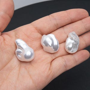 Puede incluir: Tres perlas blancas irregulares y brillantes. Las perlas tienen una superficie lisa y reflectante, con un brillo suave y nacarado. Las perlas se sostienen en una mano, sobre un fondo oscuro.