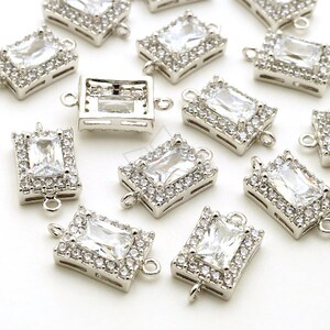 May include: Silver-toned rectangular jewelry components with a clear, rectangular center stone surrounded by smaller, clear stones. Each component has loops on the sides for attachment. The components are scattered on a white surface.