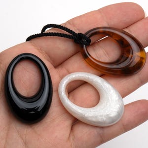 May include: Three oval pendants in black, white, and tortoiseshell brown. The black pendant has a glossy finish, the white pendant has a pearlescent sheen, and the brown pendant is attached to a black cord. These pendants are suitable for necklaces.