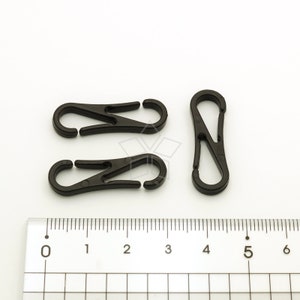 Pu&ograve; includere: Tre moschettoni in plastica nera sono disposti su una superficie bianca accanto a un righello. Due moschettoni sono aperti e uno &egrave; chiuso. Le clip sono realizzate in un materiale plastico resistente e sono progettate per vari usi.