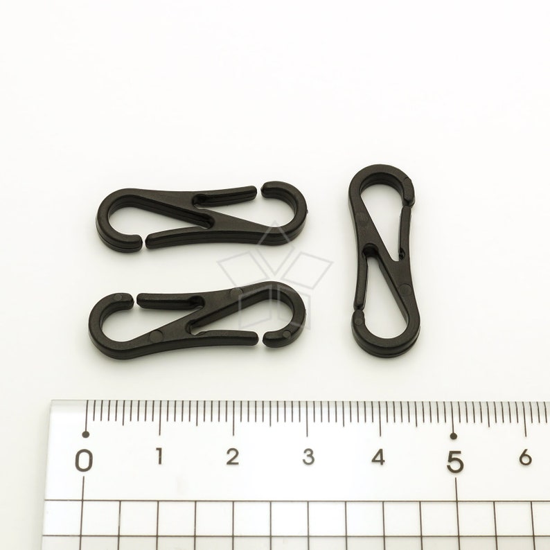 Pu&ograve; includere: Tre moschettoni in plastica nera sono disposti su una superficie bianca accanto a un righello. Due moschettoni sono aperti e uno &egrave; chiuso. Le clip sono realizzate in un materiale plastico resistente e sono progettate per vari usi.