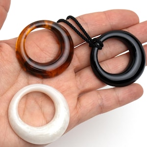 May include: Three circular pendants in shades of brown, black, and white. The brown pendant has a tortoiseshell pattern. The black pendant is attached to a black cord. The white pendant has a pearlescent sheen. Jewelry.