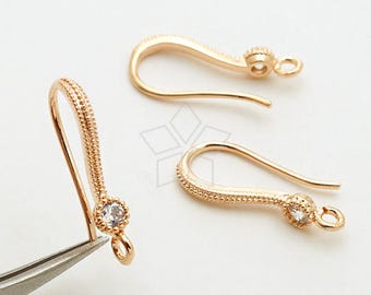May include: Close-up of three gold-colored earring hooks. Each hook has a curved design with a decorative beaded texture and a small, clear gemstone. One hook is held by tweezers.
