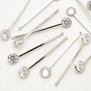 Puede incluir: Un conjunto de componentes de joyería plateados. Cada pieza presenta una barra larga y delgada con un pequeño lazo en un extremo y un cristal redondo y transparente en el otro. Los cristales brillan, reflejando la luz.