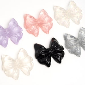 May include: Six different colored resin bows. The bows are in shades of pink, purple, white, black, and silver. The bows are all about 2 inches wide.