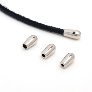 May include: Close-up of silver metal cord ends with a dark blue cord. Three cord ends are scattered, and one is attached to the cord. The cord ends are smooth, shiny, and have a small hole at the base.