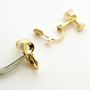 May include: Close-up of gold-colored clip-on earring findings. The findings have a small, round pad for comfort and a hinged clip mechanism. One is held by tweezers. The background is white.